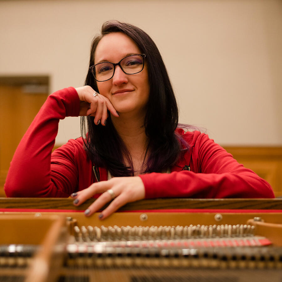 A smiling woman at the head of a piano with open strings in front. I have dark hair that has purple highlights, glasses, and a red jacket.